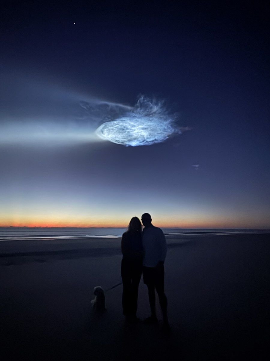 Silhouettes of a couple and their dog on Jacksonville Beach watching the glowing plume of a SpaceX launch before sunrise